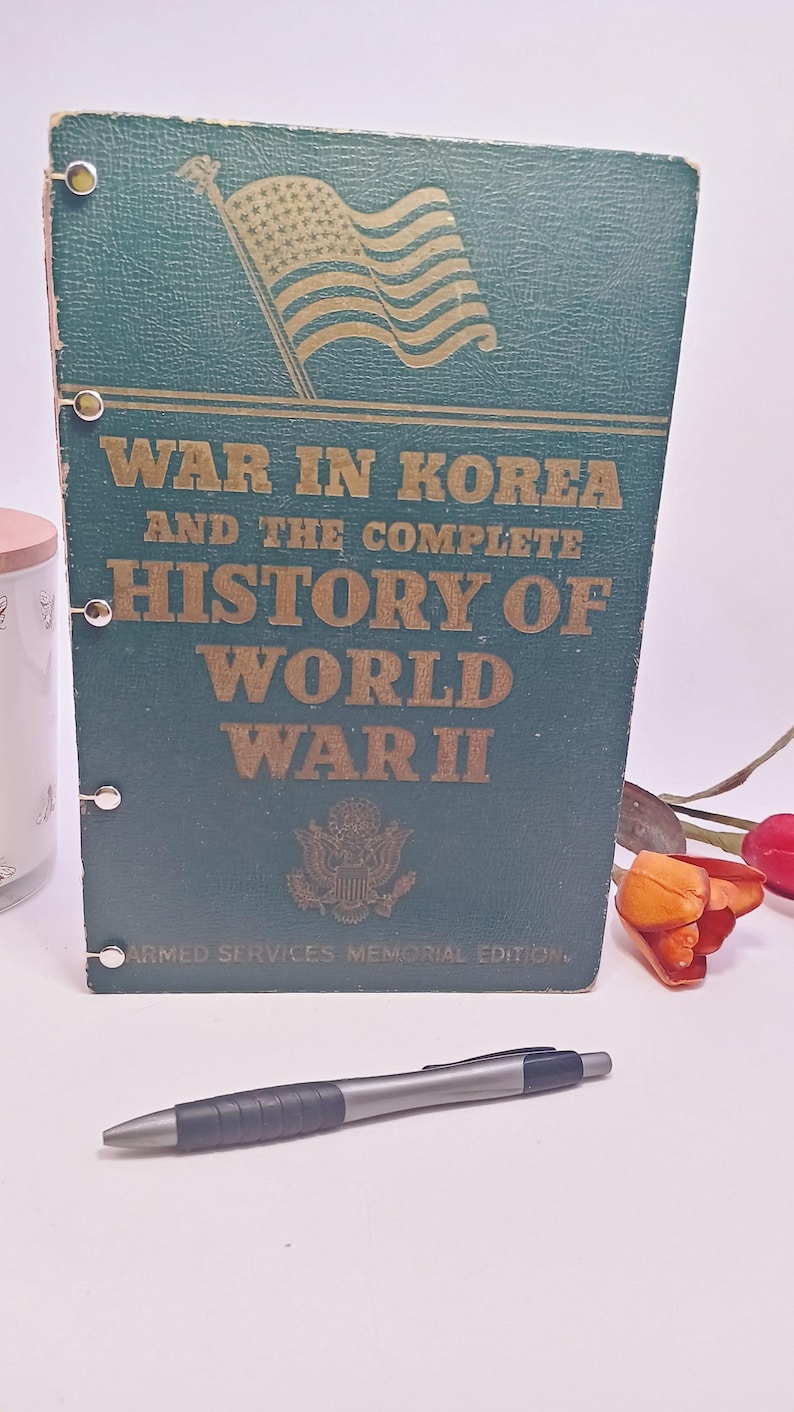 May include: A vintage book titled "War in Korea and the Complete History of World War II." The cover is dark green with gold lettering and an illustration of the American flag. A silver and black pen rests in front of the book.