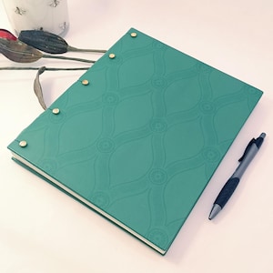May include: A teal-colored journal with a patterned cover and gold-colored binding posts. A black pen rests beside the journal. A white jar with a wooden lid and bee illustrations is in the background.
