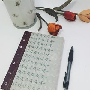 May include: A light blue candle with a wooden lid and bee illustrations, a notebook with a patterned cover, a pen, and artificial tulips. The notebook has a burgundy spine and a light blue cover with a repeating leaf design.
