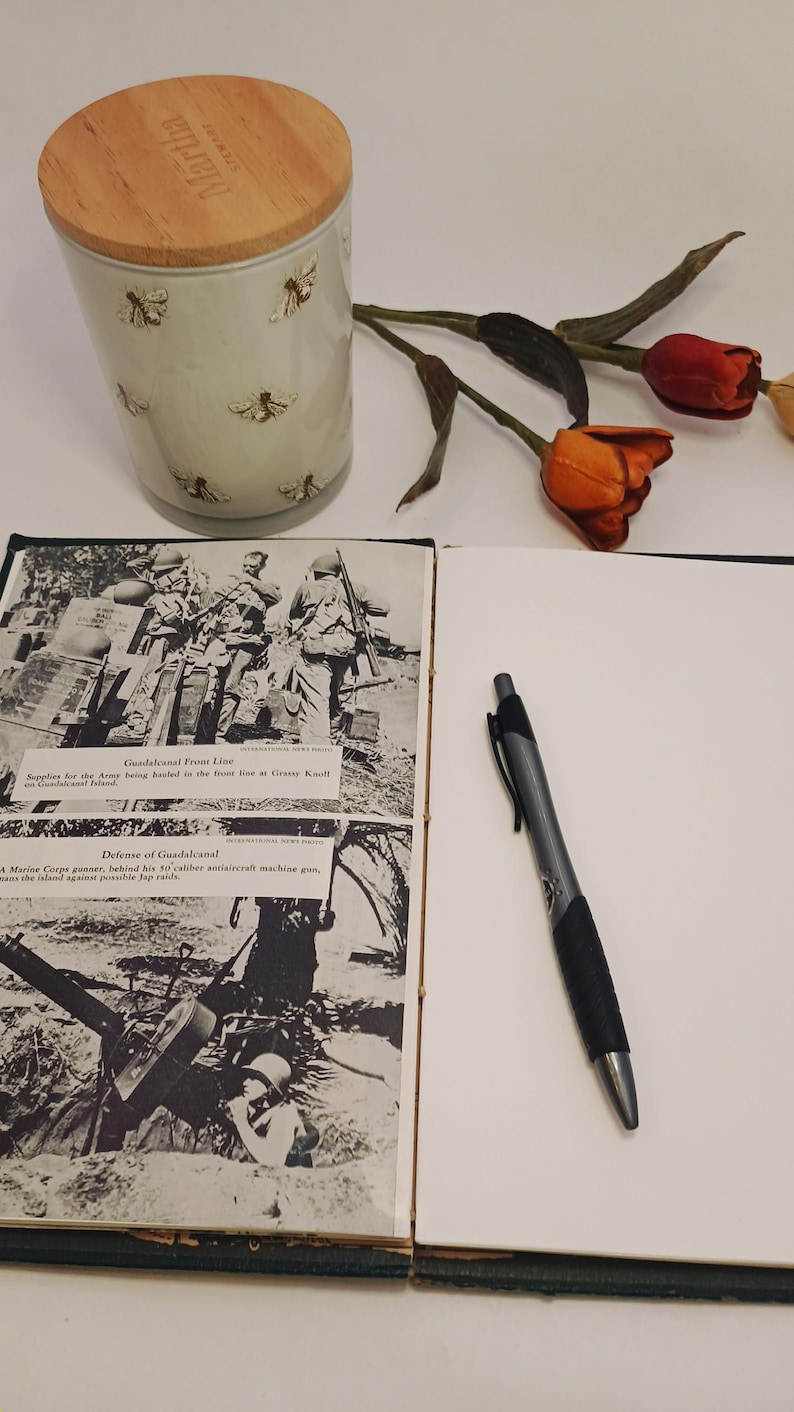 May include: A Martha Stewart ceramic jar with a wooden lid, decorated with bee motifs. A pen and a vintage book with black and white photos of soldiers are also present, along with some dried flowers.