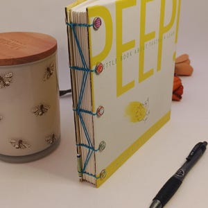 May include: A small, open book with a yellow cover and the word "DEEP" in large yellow letters. The book is bound with blue thread and decorative fasteners. A pen and a candle with bee designs are also visible.