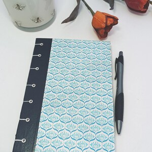 May include: A patterned journal with a blue and white design, a black spine, and a silver pen. The journal's cover features a repeating floral pattern. A decorative vase with bee illustrations and a flower are in the background.