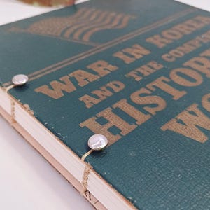 May include: A teal book titled "War in Korea and the Complete History" with gold lettering and an American flag design. Two silver fasteners secure the pages. A few colourful flowers are in the background. The book's pages are visible.