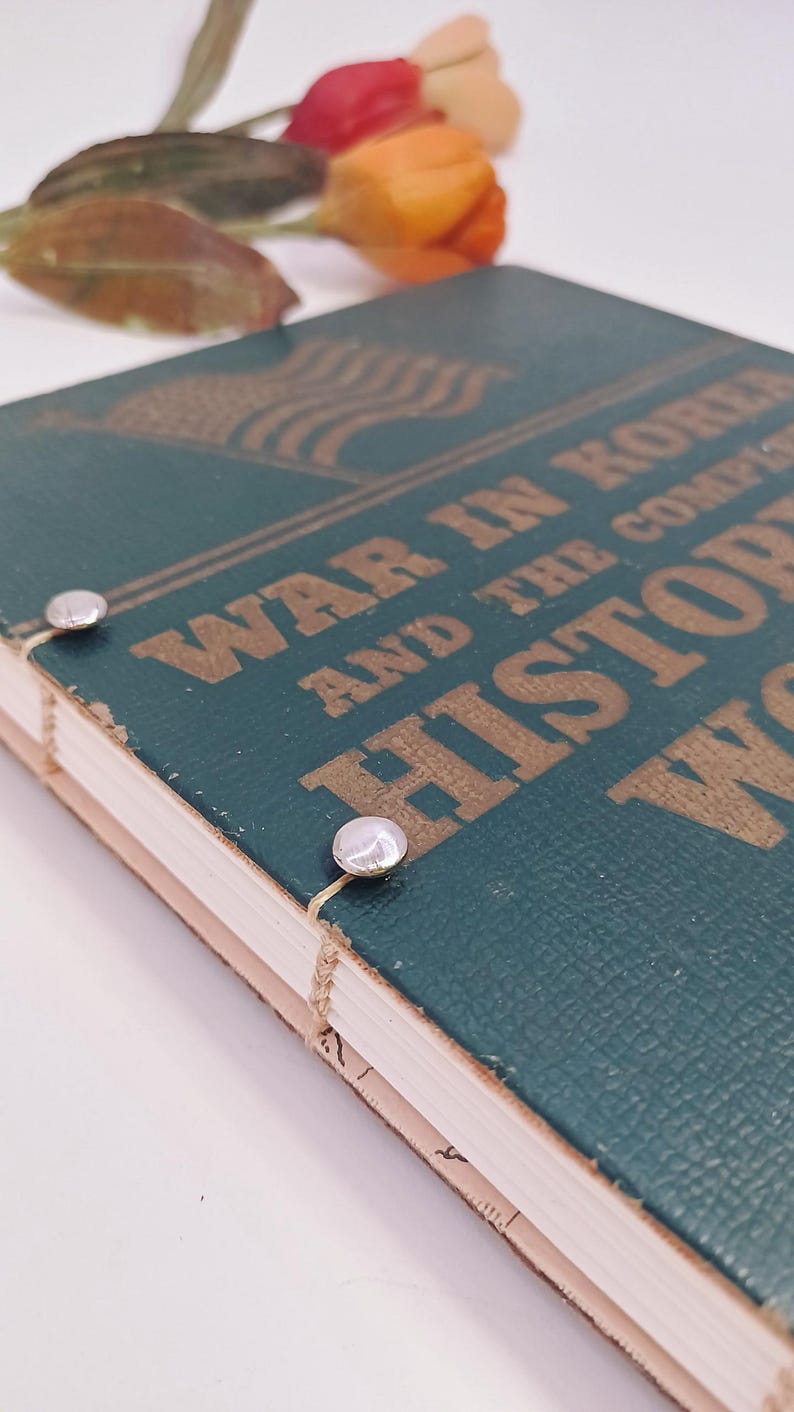 May include: A teal book titled "War in Korea and the Complete History" with gold lettering and an American flag design. Two silver fasteners secure the pages. A few colourful flowers are in the background. The book's pages are visible.