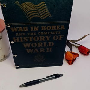 May include: A dark green book titled "WAR IN KOREA and the complete HISTORY OF WORLD WAR II" with a gold American flag graphic. A black and silver pen rests in front of the book. Red and orange tulips are in the background.