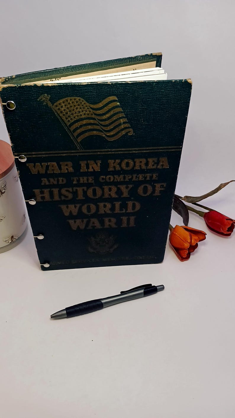 May include: A dark green book titled "WAR IN KOREA and the complete HISTORY OF WORLD WAR II" with a gold American flag graphic. A black and silver pen rests in front of the book. Red and orange tulips are in the background.