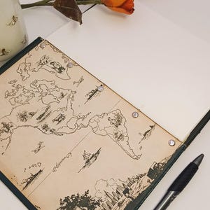 May include: An open notebook featuring a vintage world map design, a black pen, and a sprig of orange and red tulips. The notebook has a dark green border and is open to a blank page.