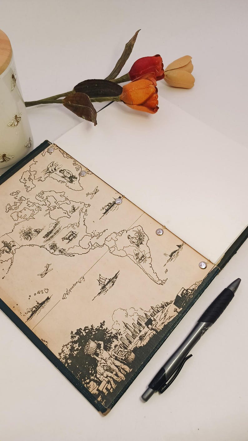 May include: An open notebook featuring a vintage world map design, a black pen, and a sprig of orange and red tulips. The notebook has a dark green border and is open to a blank page.