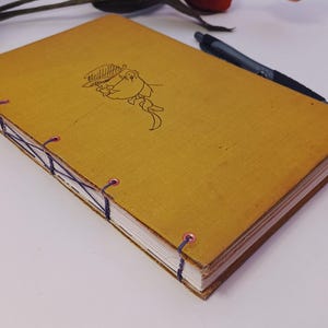 May include: Mustard-yellow journal with a hand-stitched binding. The cover features a simple illustration in black ink. A black pen rests on the journal, and decorative flowers are in the background.