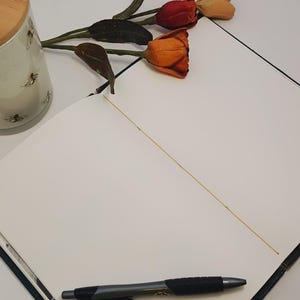 May include: An open notebook with a black pen on the page. A decorative candle with a wooden lid and bee design is visible, along with a sprig of orange and red tulips. The notebook is open to a blank page.