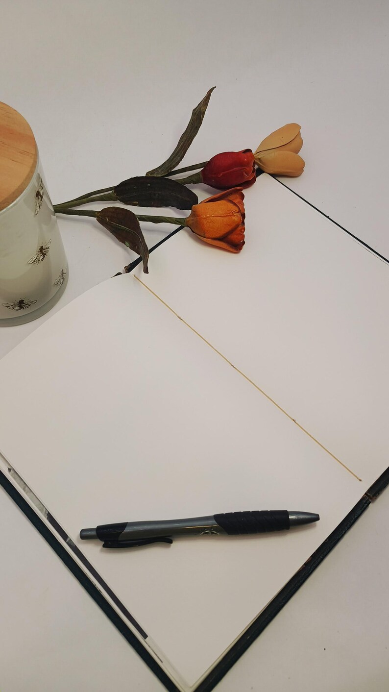 May include: An open notebook with a black pen on the page. A decorative candle with a wooden lid and bee design is visible, along with a sprig of orange and red tulips. The notebook is open to a blank page.