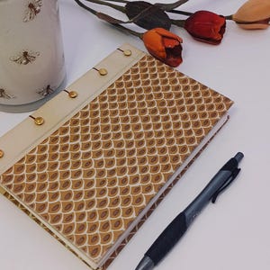 May include: A decorative journal with a brown and cream-colored patterned cover, gold accents, and a matching pen. A white candle with bee illustrations and faux tulips are also present.