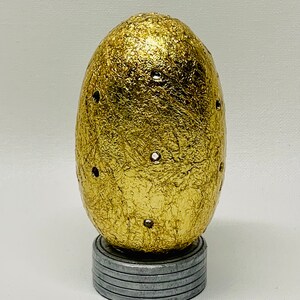 May include: A gold egg-shaped decorative object with a textured surface and small holes. It is sitting on a silver base.