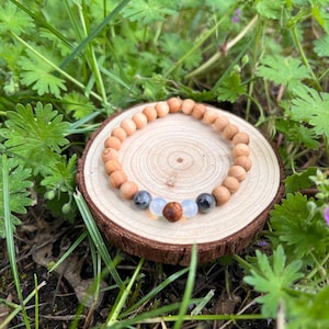 Könnte beinhalten: Ein Holzperlenarmband mit einer Mischung aus hellbraunen und farbigen Akzentperlen. Das Armband wird auf einer kleinen, runden Holzscheibe vor einem Hintergrund aus grünem Laub präsentiert.