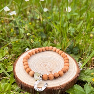 Könnte beinhalten: Ein Armband aus Holzperlen mit einem silbernen Hufeisen-Anhänger und einem grünen Herz-Anhänger. Das Armband liegt auf einer Holzscheibe und ist von grünem Gras und weißen Blumen umgeben.