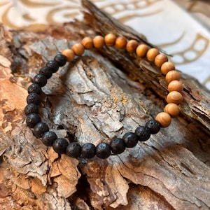 Könnte beinhalten: Ein Perlenarmband mit einem abwechselnden Muster aus glatten, hellbraunen Holzperlen und texturierten, porösen schwarzen Lavasteinperlen. Das kreisförmige Accessoire ist auf einem flexiblen Band aufgezogen und wechselt zwischen Holz- und Steinabschnitten.