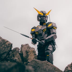 Puede incluir: Una persona con un traje de robot negro y dorado está de pie sobre una superficie rocosa, sosteniendo un arma larga y delgada. El traje tiene un casco con cuernos amarillos y detalles en forma de alas en los hombros. El fondo es un cielo nublado.