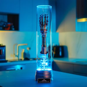 Puede incluir: Una mano robótica encerrada en un cilindro de vidrio transparente sobre una base plateada con el texto "Cyberdyne Systems" en la parte delantera. La mano está hecha de metal y tiene un acabado plateado. El cilindro está iluminado desde abajo con una luz azul.