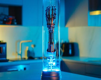 May include: A robotic hand encased in a clear glass cylinder on a silver base with the text "Cyberdyne Systems" on the front. The hand is made of metal and has a silver finish. The cylinder is illuminated from below with a blue light.