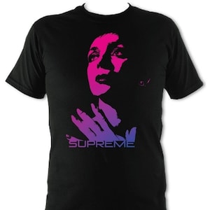 Op de afbeelding: Zwart T-shirt met een roze en blauwe gradi&euml;nt afbeelding van een gezicht en handen van een persoon met het woord "SUPREME" eronder gedrukt.