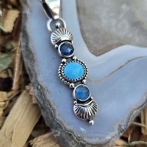 May include: A silver pendant necklace featuring a vertical arrangement of three circular blue gemstones. The design includes a dark blue stone at the top, a bright turquoise stone in the center, and a light blue stone at the bottom, accented by silver metalwork with fan-shaped details.