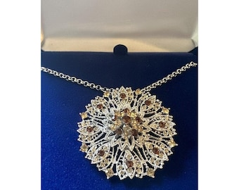 May include: A silver necklace with a large, ornate pendant featuring a floral design with brown and clear crystals. The pendant is in the shape of a flower with a center of clear crystals surrounded by brown crystals. The necklace is in a blue box with the words "Camrose & Kross" printed on the top.