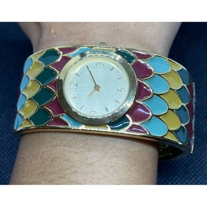 May include: A gold-tone bangle watch with a white face and black hands. The watch band is decorated with a colorful mosaic pattern of green, blue, yellow, and red.
