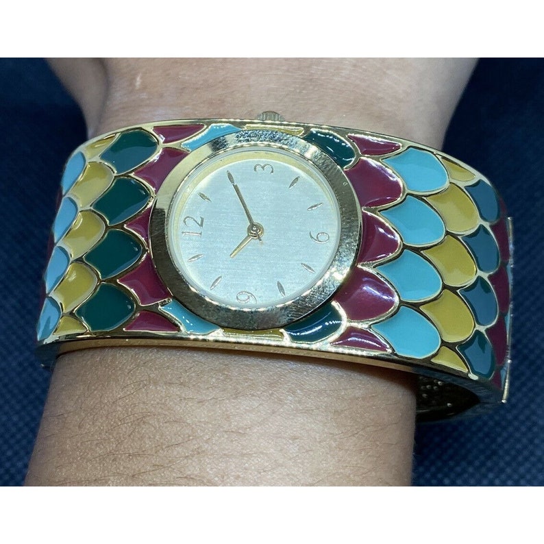 May include: A gold-tone bangle watch with a white face and black hands. The watch band is decorated with a colorful mosaic pattern of green, blue, yellow, and red.