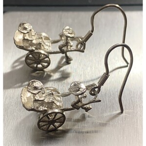 May include: A pair of silver earrings featuring a detailed miniature rickshaw with a rider. The earrings have a simple hook closure.