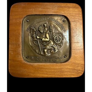 Può includere: Una targa in legno con un rilievo in ottone di San Cristoforo che porta un bambino. La targa ha il testo "Behold Saint Christopher" e "Then go your way in safety".