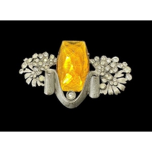 Puede incluir: Un broche de plata con una gran piedra de vidrio facetada de color ámbar en el centro. La piedra está rodeada por un diseño de plata que se asemeja a una flor estilizada. El broche está decorado con piedras de vidrio más pequeñas y transparentes.