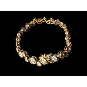 May include: A gold-toned bracelet with multiple clear, round gemstones set in individual bezels. The bracelet has a secure clasp and is displayed against a black background. The jewelry piece is likely made of gold-plated metal.