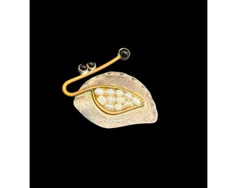 Könnte beinhalten: Eine einzigartige Brosche mit blattartigem Design. Die Brosche hat eine silberfarbene Basis mit goldfarbener Kontur und ist mit kleinen, weißen Perlen gefüllt. Ein gebogener goldfarbener Stiel erstreckt sich vom Blatt und endet in drei schwarzen Kugeln.