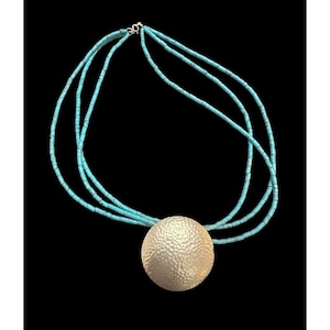 May include: A turquoise beaded necklace with a large, hammered silver pendant. The necklace features three strands of small, turquoise-colored beads. The pendant is a large, textured silver disc. The necklace is displayed against a black background.