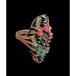 May include: Silver-toned ring with an oval cluster of gemstones. The ring features red, green, black, and yellow stones. The band has a split design. The ring is set against a black background.
