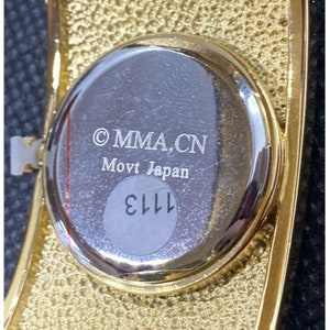 May include: The back of a gold-toned watch with a silver face. The watch face has the text "&copy; MMA.CN Movt Japan" and the number "1113" printed on it.