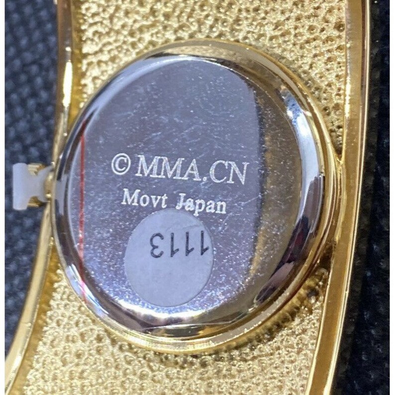 May include: The back of a gold-toned watch with a silver face. The watch face has the text "&copy; MMA.CN Movt Japan" and the number "1113" printed on it.