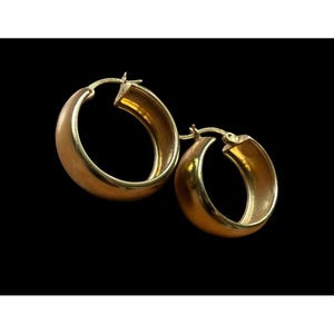 May include: A pair of gold-colored hoop earrings. The earrings have a thick, rounded design and a secure clasp. The interior of the hoops has a slightly lighter gold tone. The background is solid black.