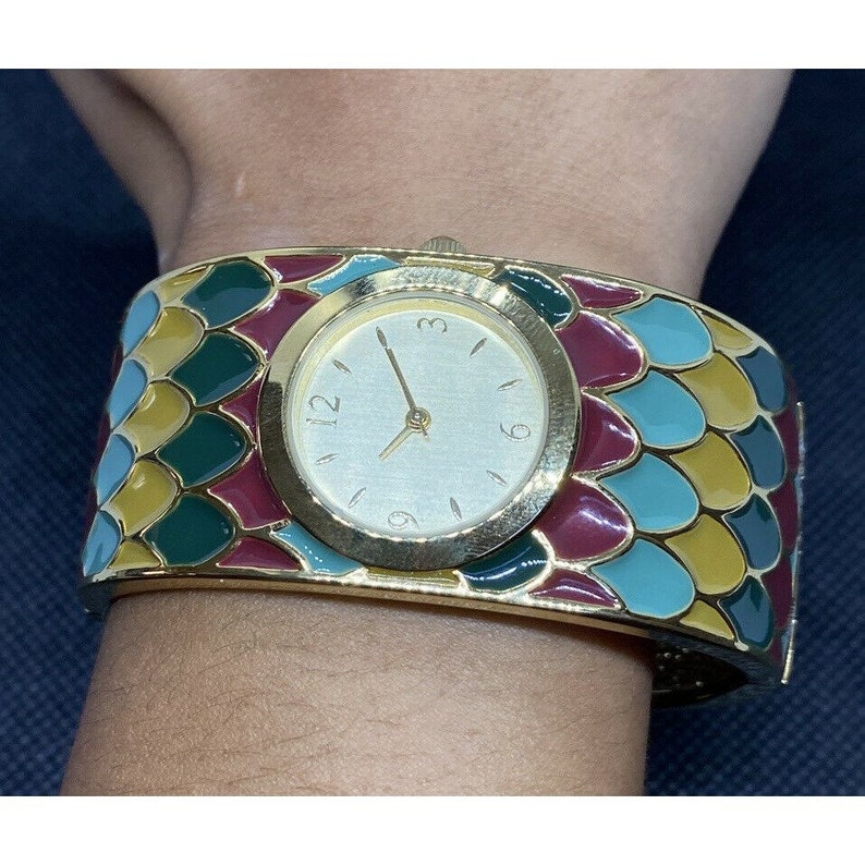 May include: A gold-tone watch with a white face and black hour and minute hands. The watch band is a wide cuff with a colorful, mosaic-like design featuring green, blue, yellow, and red.