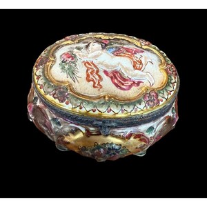 May include: A vintage porcelain trinket box with a detailed hand-painted design of a cherub with wings and a floral motif. The box has a gold-gilded trim and a lid with a hinged closure.