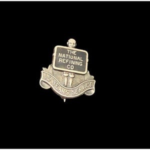 May include: A gold-coloured pin with a black sign that reads "The National Refining Co." and a figure holding the sign. The pin also has a banner that reads "15 Years Service".
