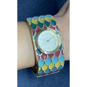 May include: A gold-tone bangle watch with a white face and black hands. The watch band is decorated with a colorful pattern of green, blue, yellow, and red scales.