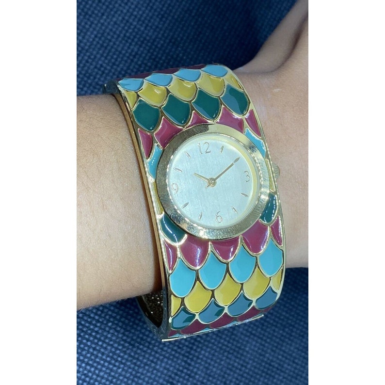 May include: A gold-tone bangle watch with a white face and black hands. The watch band is decorated with a colorful pattern of green, blue, yellow, and red scales.