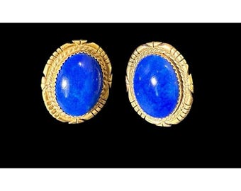 Könnte beinhalten: Ein Paar goldfarbene ovale Ohrringe mit einem leuchtend blauen Mittelstein. Die Ohrringe haben einen detaillierten, strukturierten Rand. Der Hintergrund ist schwarz und hebt den Schmuck hervor.