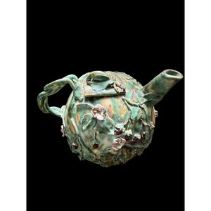 Puede incluir: Tetera de cerámica artesanal con forma esférica, adornada con hojas verdes, flores rosas y pequeñas bayas rojas. El asa está torcida y el pico curvado. La tetera tiene un acabado mate y está sobre un fondo negro.