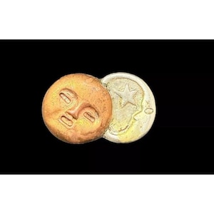 May include: A two-part metal pendant featuring a stylized face on one side and a star on the other. The face is copper-colored and the star is silver-colored.