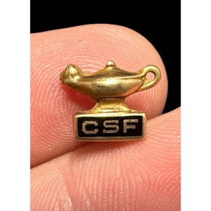 May include: Gold-colored pin with a small oil lamp design and the letters "CSF" on a black background.