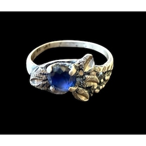 May include: A silver ring with a large, round, deep blue gemstone at the center. The ring has an ornate, textured design around the gemstone and along the band. The ring is set against a black background.