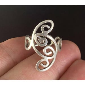May include: A silver-colored ring with an openwork design featuring swirling, abstract shapes. The ring has a band and a decorative top with a series of curves and loops. The ring is shown against a dark background.