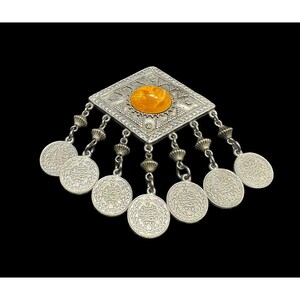 Może przedstawiać: Srebrny wisiorek z kwadratową ramą i bursztynowym kamieniem w środku. Rama jest ozdobiona misternymi wzorami, a wisiorek wisi na łańcuszku z kilkoma małymi srebrnymi monetami.